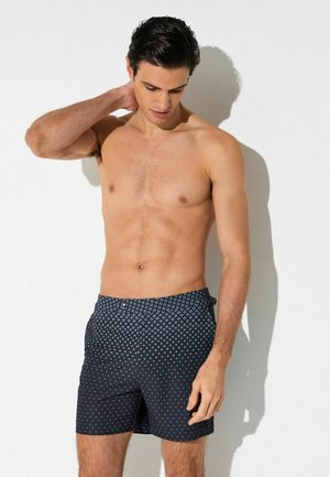 Mężczyzna bez koszulki o ciemnych włosach, ubrany w granatowe spodenki kąpielowe z białymi geometrycznymi wzorami, stojący na tle białej ściany.