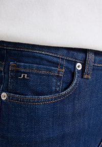 Tmavě modré džíny z denimu s oranžovým prošíváním, které mají malou přední kapsu a kovové zapínání na knoflík v pase.