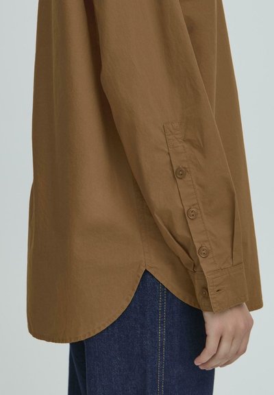 Bruine, langslavige shirt met een ronde zoom, knoopdetails op de mouw en een gladde textuur, gecombineerd met een donkerdenim jeans.