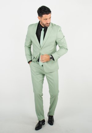 Un costume vert clair avec une chemise noire et une cravate assortie, complété par une montre. Le tissu semble lisse, et les chaussures sont noires et brillantes.