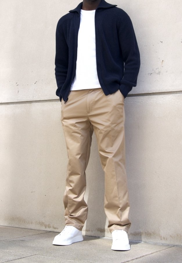 Homme portant un pantalon beige, des baskets blanches, un t-shirt blanc et un pull zippé bleu marine, debout contre un mur clair.