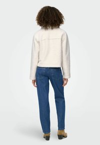 Persona con capelli ricci che indossa un giubbotto bianco soffice, jeans blu e stivali con tacco color tan, in piedi di spalle su uno sfondo neutro.