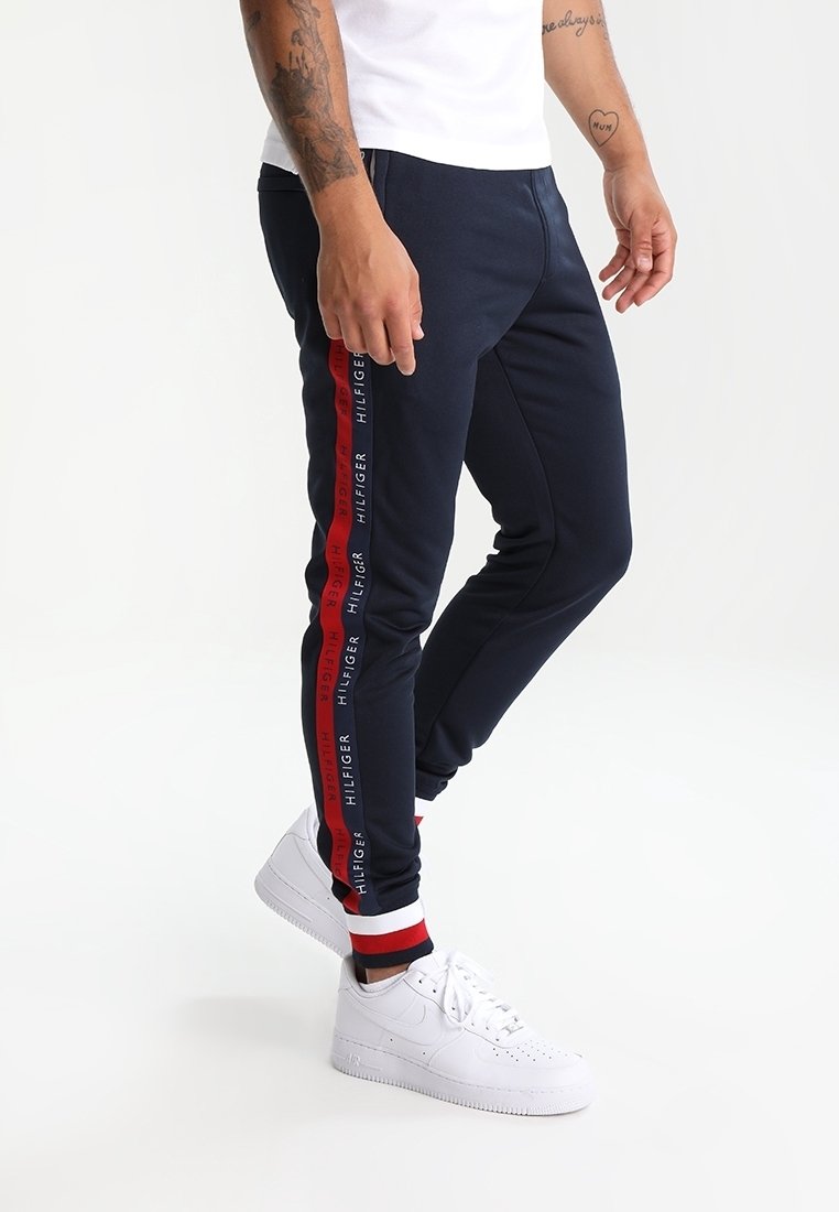 Marineblaue Jogginghosen mit roten und weißen Seitenstreifen, auf denen der Text "HILFIGER" steht. Hergestellt aus einem glatten Stoff mit elastischen Bündchen, kombiniert mit weißen Turnschuhen.