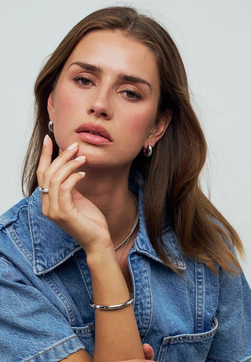 Silver smycken på en modell med medelbrunt hår. Hon bär hoopörhängen och lager med halsband, klädd i en denimskjorta. Händerna visar en ring och ett armband.