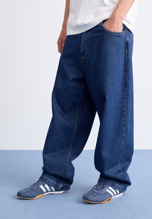 Baggy blue denim jeans with high waist, side pockets, and contrast stitching, paired with gray sneakers featuring white stripes.