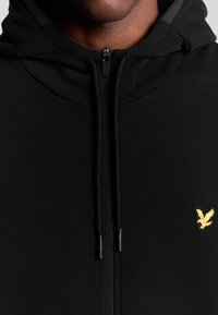 Felpa nera con zip, caratterizzata da una finitura texturizzata, con un logo dorato a forma di aquila sul petto e dettagli con coulisse sul cappuccio.
