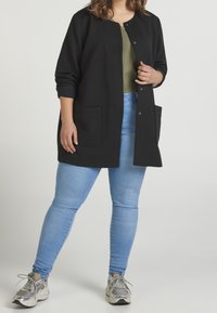 Czarna, prążkowana długa kurtka z zatrzaskami i przednimi kieszeniami, noszona na zielonym topie i zestawiona z jasnoniebieskimi obcisłymi dżinsami oraz srebrnymi tenisówkami.