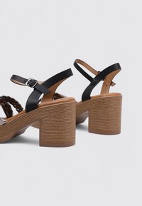 Sandalias de tacón de cuero negro con un diseño trenzado, una plataforma gruesa marrón y correas de tobillo ajustables. Suela texturizada para mayor agarre.