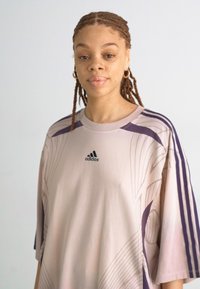 Svijetlopurple oversized majica s tamnopurple detaljima, na kojoj je Adidas logo, uzorci zakrivljenih linija i kratki rukavi.
