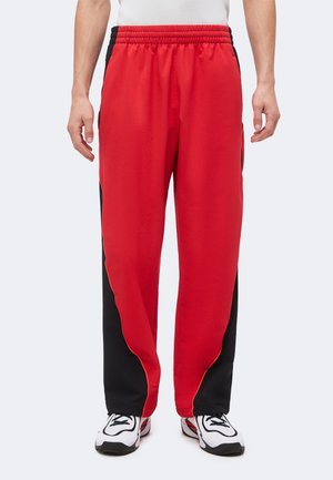 Homme portant un pantalon de sport rouge et noir avec une ceinture élastique et des baskets blanches, debout devant un fond uni.