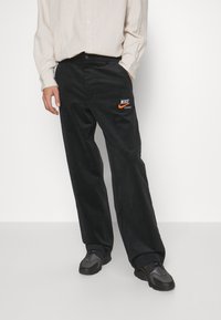 Svarta kordfløjlsbyxor med en avslappnad passform, med en broderad orange Nike-logotyp på vänster ficka. Bärs tillsammans med en beige skjorta med knappar.