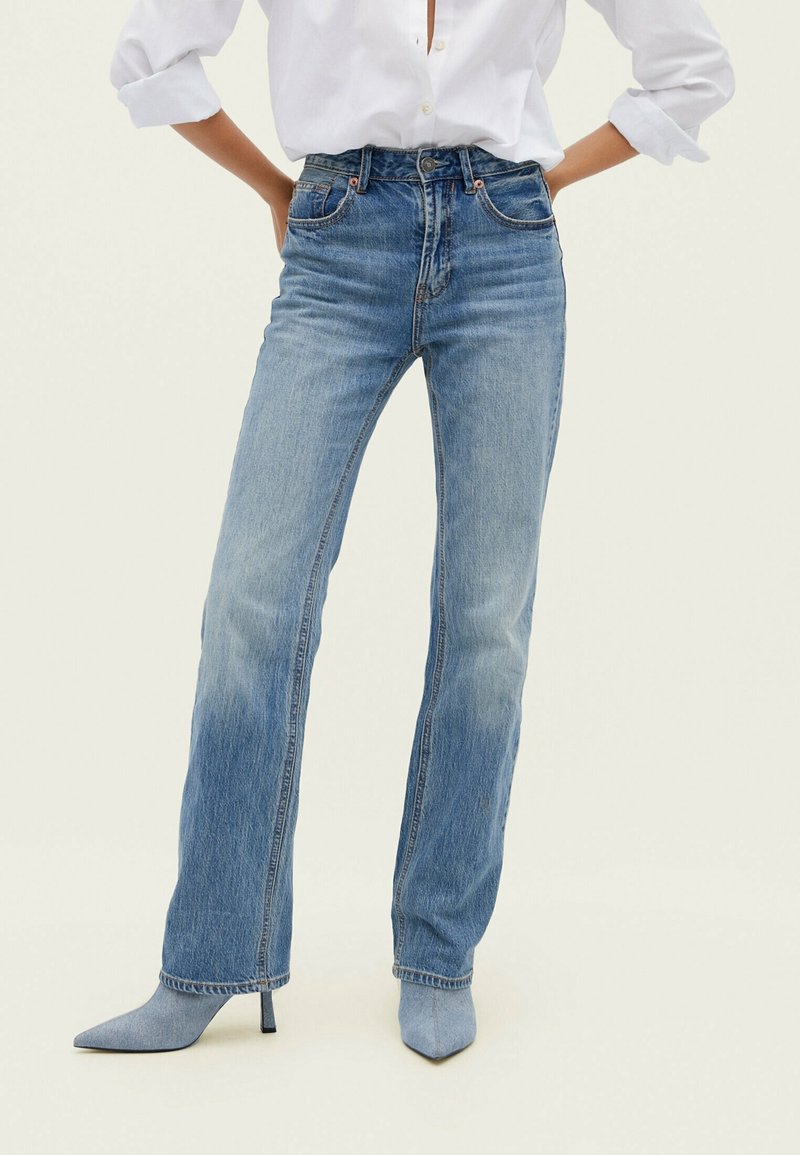 Person wearing two-tone blue jeans with a lighter wash on the inner legs, paired with a white shirt and light blue pointed-toe heeled boots.