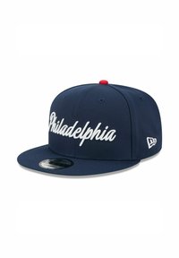Casquette snapback bleu marine avec inscription blanche "Philadelphia" en écriture cursive. Elle présente une visière plate et un bouton rouge sur le dessus, avec un détail de logo sur le côté.