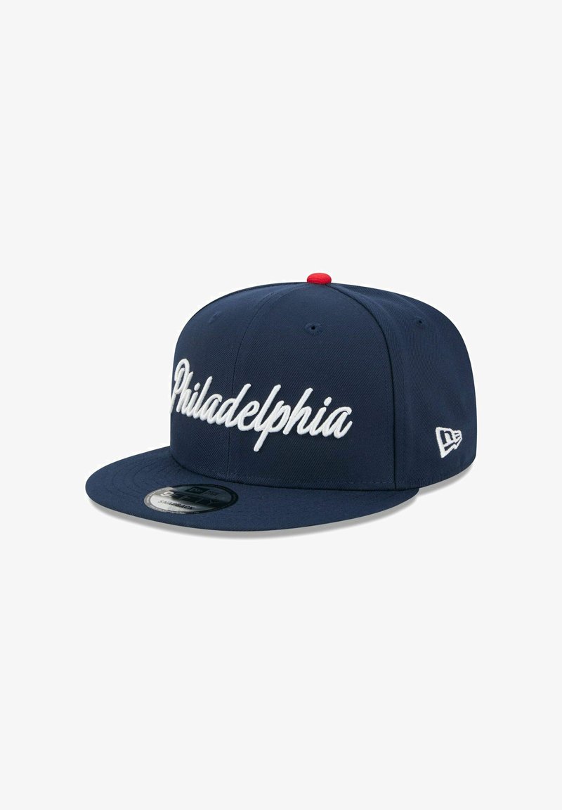 Casquette snapback bleu marine avec inscription blanche "Philadelphia" en écriture cursive. Elle présente une visière plate et un bouton rouge sur le dessus, avec un détail de logo sur le côté.