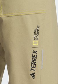 Beige tygbyxor med gula sömmar, med svart och gul logotyp från "Terrex" och "National Geographic." Inkluderar en sidoficka.
