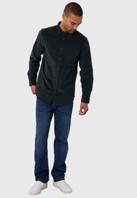 Mørkegrøn skjorte med knapper og en brystlomme, båret med blå jeans og hvide sneakers. Stoffet ser struktureret og blødt ud.