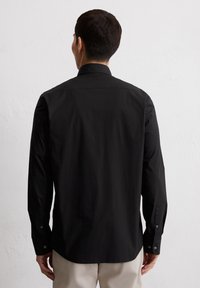 Camisetta nera a maniche lunghe con colletto appuntito, polsini con bottone e una texture liscia. Presenta una leggera lucentezza e una vestibilità sartoriale.