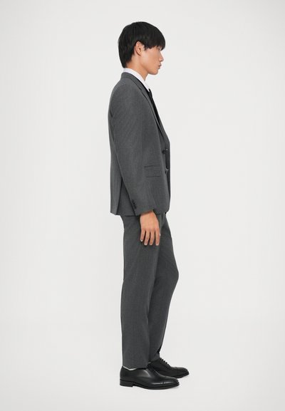 Traje gris a medida con un corte ajustado, que presenta una chaqueta de un solo botón y solapas muesca, combinado con zapatos de vestir negros.