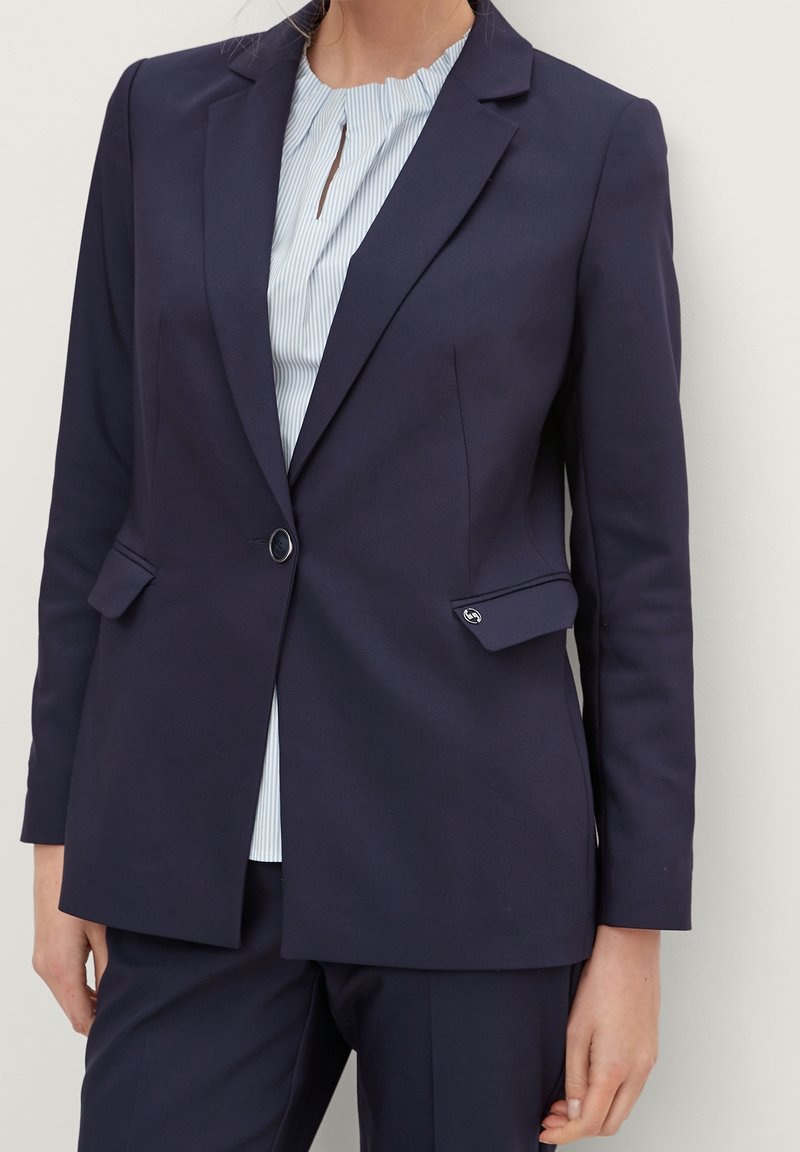 Blazer azul marino con un diseño entallado, solapas de muesca, dos bolsillos frontales y cierre de un solo botón, combinado con una blusa de rayas clara.