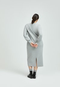 Langärmliges graues Kleid mit einem Rückenschlitz, leicht strukturiertem Stoff, taillierter Silhouette und kontrastierenden dunkel gefärbten Schnürstiefeletten.