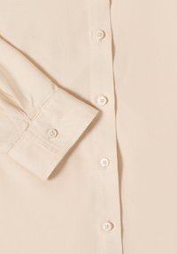 Poignet de chemise beige à manches longues avec détail boutonné et une rangée verticale de boutons blancs sur un fond de tissu lisse.