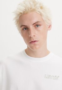 Camiseta de algodón blanca con una textura suave y un sutil logotipo gris "LEVI'S" en el lado izquierdo del pecho. Cuello redondo, corte relajado.