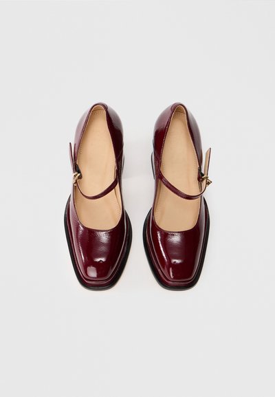 Μπορντώ παπούτσια από πατεντέ δέρμα με τετράγωνη μύτη, κομμένες πλευρές και ρυθμιζόμενο λουρί με χρυσή πόρπη. Σόλες επίπεδες.