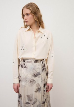 Giovane donna che indossa una camicia color crema con ricami floreali e una gonna a vita alta con motivi astratti neri e grigi.