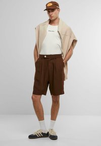 Braune, maßgeschneiderte Shorts mit hoher Taille und Vorderfalten, kombiniert mit einem hellbeigen Schal und einem weißen Shirt, abgerundet mit sportlichen Schuhen und einer Kappe.