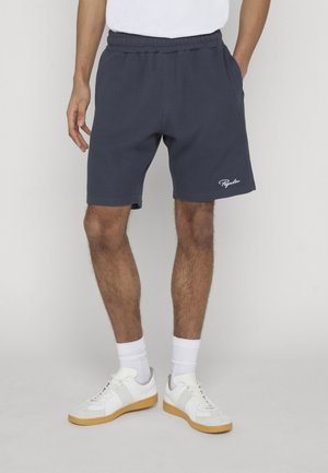 Man wearing navy blue ribbed shorts with white logo, white socks, and white sneakers with gum soles, standing with hands in pockets.