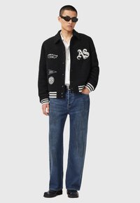 Mand iført sort letterman-jakke med lapper, hvid skjorte, blå jeans, sorte sko og sorte solbriller, der står mod en grå baggrund.