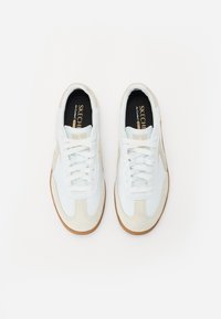 Hvit sneakers med beige semsket skinn-detaljer, snøring, gummisåle, og svarte innersåler med merkevarenavn. Jevn tekstur gjennom hele.