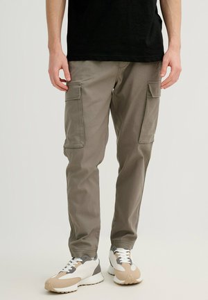 Persona con pantalones cargo grises con bolsillos laterales y zapatillas blancas con detalles beige y grises, de pie frente a un fondo neutro.