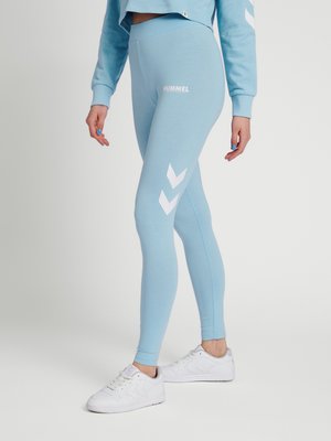 Person iført lyseblå Hummel leggings med hvide chevron-logoer på benene og hvide sneakers, stående mod en ensfarvet lys baggrund.