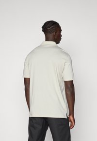Polo à manches courtes en tissu beige clair avec un col classique, des fentes sur les côtés et une texture douce. Vu de dos.