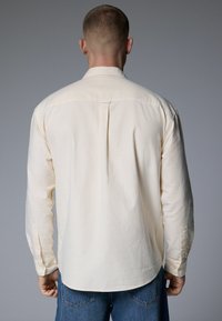 Light beige shirt with thin vertical stripes, long sleeves, buttoned cuffs, and a yoke detail at the back. Smooth fabric texture.