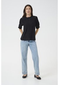 Blusa nera semi-trasparente con maniche a sbuffo e scollatura ondulata, abbinata a jeans azzurri a vestibilità rilassata e ballerine nere.