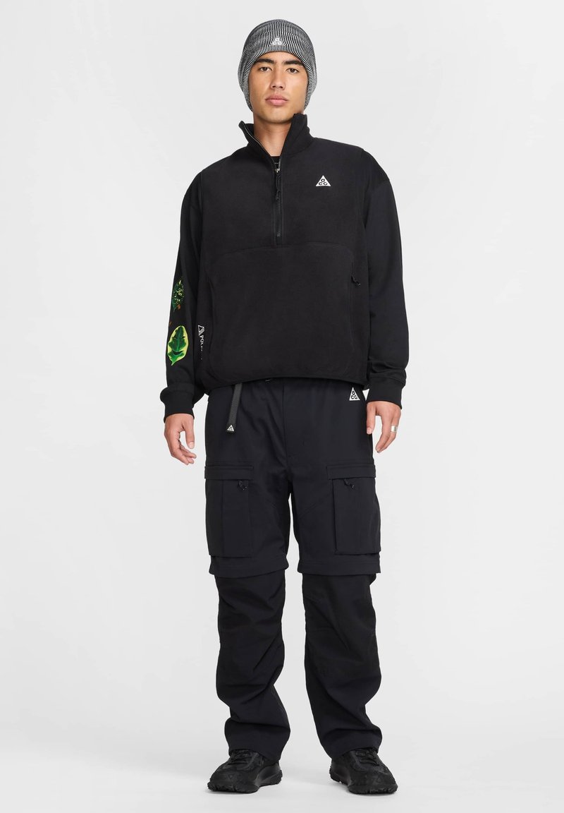Black fleece pullover with a zip neckline and green leaf pattern on the sleeve, paired with black cargo pants featuring multiple pockets.