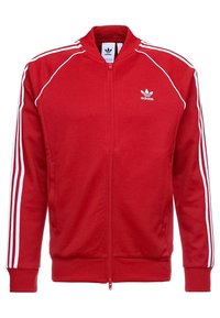 Punainen vetskari takki, joka on valmistettu pehmeästä kankaasta. Takissa on valkoinen kolme viivaa -design olkapäillä ja hihoissa, sekä Adidas-logo vasemmassa rinnassa.