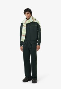 Groene trainingspak gemaakt van zachte stof met gele piping, gecombineerd met een gestreepte zachte sjaal in groen, blauw en roze. Model staat met één hand op de heup.