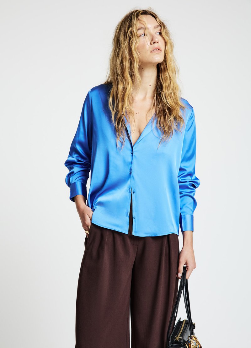 Camicia in raso blu vivace con scollo a V e maniche lunghe, abbinata a pantaloni marroni a gamba larga e una borsa nera.