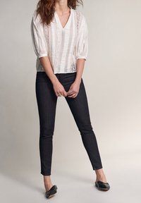 Vit blus med V-ringning, struktur tyg med vertikala ränder och spetsdetaljer, kombinerad med mörka tajta jeans och svarta loafers.