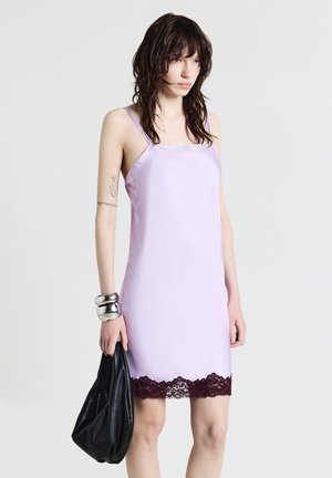 Lila Satin-Kleid mit dünnen Trägern und einem bordeauxroten Spitzenrand. Das Model hält eine schwarze Ledertasche und trägt ein massives metallisches Armband.