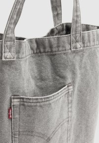 Sac fourre-tout en toile grise avec deux poignées robustes, doté d'une poche avant avec des coutures blanches et une petite étiquette de marque rouge.