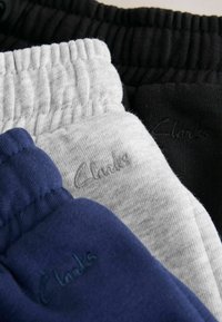 Bandes élastiques de pantalons de survêtement bleus, gris et noirs pliés et empilés, chacun avec un discret logo "Clarks" brodé sur le tissu.