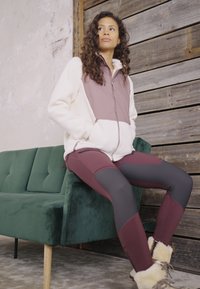 Veste en polaire zippée crème et mauve, associée à un legging rouge foncé, assise sur un canapé en velours vert contre un fond en bois.