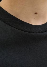 Crna pamučna majica s rebrastim ovratnikom. Prikaz u blizini pokazuje glatku teksturu i suptilne detalje u tkanju. Bez uzoraka ili ukrasa.