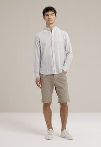 Pruhovaná košeľa s button-up v svetlosivej farbe, skombinovaná s béžovými šortkami. Model má na sebe biele tenisky a stojí pred neutrálnym pozadím.