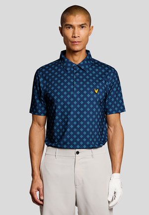Homme portant un polo bleu marine à motifs avec un logo jaune, un pantalon beige et un gant de golf blanc, debout devant un fond gris clair.