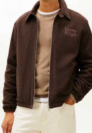 Mann trägt eine dunkelbraune Zip-Jacke mit "Tommy Hilfiger"-Logo auf der Brust über einem beigen Pullover und weißen Hosen.
